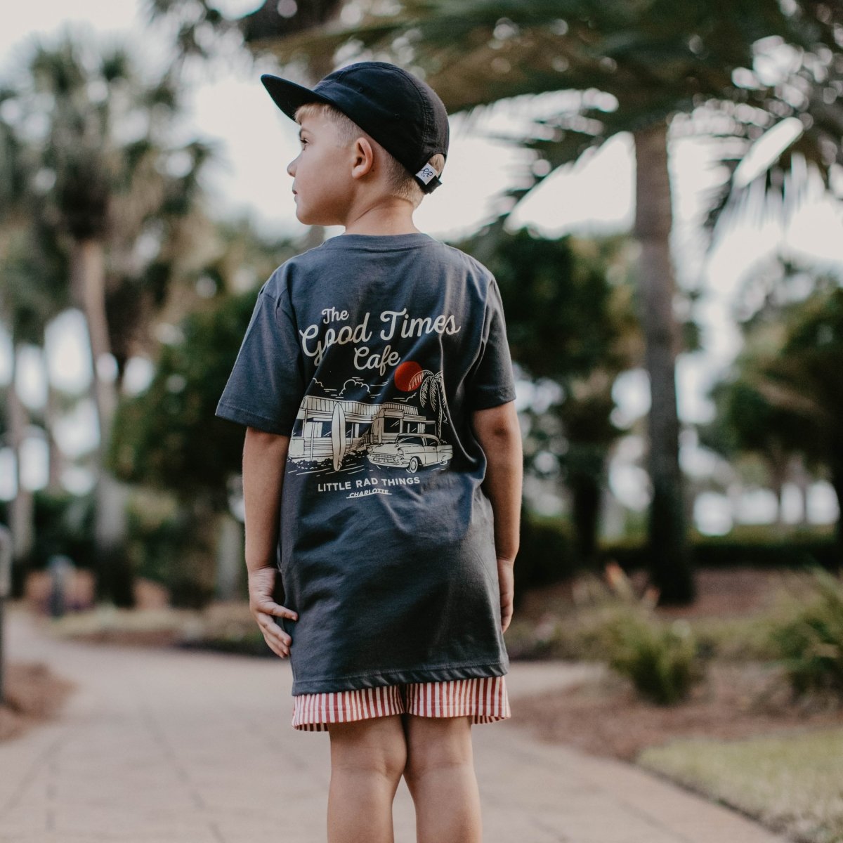 Back view of a young boy wearing a backward baseball cap and a black organic cotton graphic t-shirt outside.